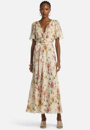 Frau trägt ein langes, florales, cremefarbenes Kleid mit kurzen, flatternden Ärmeln, V-Ausschnitt, einem Taillengürtel und strukturierte, offene Absätze.