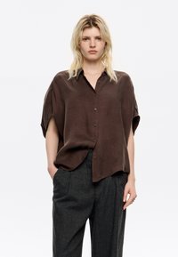 Brown, short-sleeve, button-up shirt made of soft, lightweight fabric with a relaxed fit and subtle sheen, paired with gray tailored trousers.