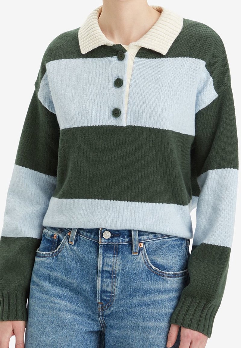 Femme portant un pull en maille rayé vert foncé et bleu clair avec un col côtelé crème et trois boutons foncés, associé à un jean bleu.