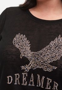 Černé tričko s krátkým rukávem s designem orla vyrobeným z rhinestones a textem "DREAMER", má kulatý výstřih a jemnou texturu.