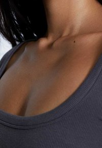 Close-up van de nek, sleutelbeen en bovenste borst van een vrouw, die een donker geribbeld tanktopje draagt, met kort zwart haar zichtbaar.