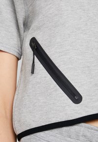 Šedá mikina s krátkým rukávem a černou kapsou na zip. Látka je měkká s jemnou strukturou, na lemu je černý lem.