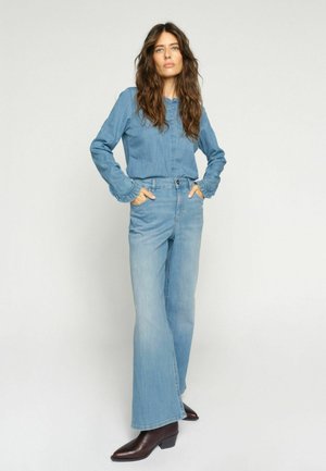 Žena s dlhými vlnitými vlasmi, oblečená v modrej rifľovej košeli a rozšírených džínsoch, stojaca s rukami v priečinkách a hnedými členkovými čižmami.