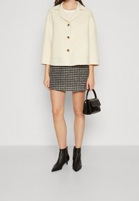 Crème wollen gemêleerde cropcoat met gouden knopen, zwart-wit houndstooth minirok, zwarte enkellaarsjes en een kleine gestructureerde handtas.