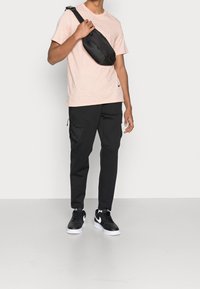 Young man wearing pale pink t-shirt, black cargo pants, black and white sneakers, and a black crossbody bag.