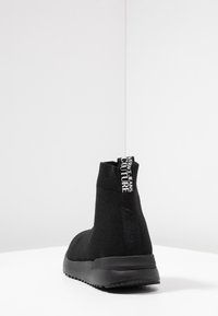 Black high-top sneaker made from stretchy fabric with a textured feel, featuring a pull tab with white logo and a thick black sole.