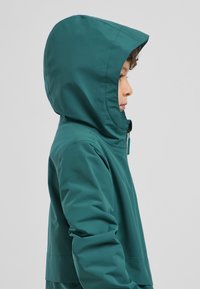 Giacca impermeabile teal con cappuccio, dotata di zip frontale e polsini. Texture liscia con finitura opaca, progettata per una vestibilità aderente.