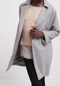 Ljusgrå, skräddarsydd kappa med lång, avslappnad passform och slagkrage. Bärs över en beige strukturerad tröja och svarta byxor.