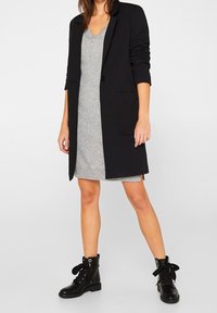 Blazer nero con vestibilità su misura, indossato sopra un vestito grigio con scollo a V. Stivaletti neri con lacci e occhielli completano il look.