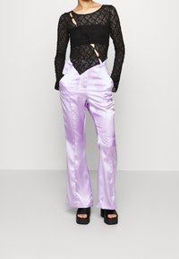 Black lace long-sleeve top with cut-out details, paired with high-waisted lilac satin trousers. Features wide legs and a shiny texture.