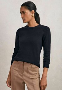 Donna dai capelli scuri raccolti in una coda di cavallo, che indossa orecchini d'oro, un maglione nero a maniche lunghe e pantaloni beige con tasche a toppa frontali.