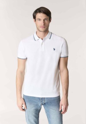 Uomo che indossa una polo bianca con rifiniture blu navy e logo blu, abbinata a jeans azzurri, in piedi davanti a uno sfondo chiaro.