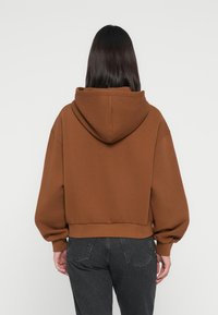 Brun hettegenser med en cropped design, ribbestrikket hem og vide ermer. Båret med mørkeblå jeans, sett fra baksiden.
