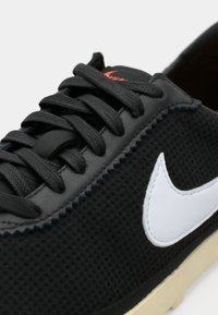 Μαύρο αθλητικό παπούτσι με υφασμάτινη επένδυση σε υφή, λευκό λογότυπο σούσι και αντίθετη μπεζ σόλα. Διαθέτει μαύρες κορδόνες και ενισχυμένες ραφές.