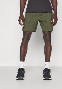 Shorts vert olive avec une finition texturée, présentant des détails de couture visibles, associés à des chaussures de sport noires et des chaussettes blanches.