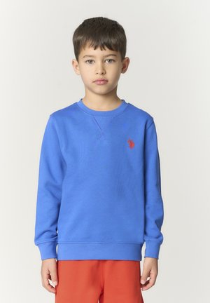 Ragazzo con capelli corti e scuri che indossa una felpa blu con logo rosso e pantaloni rossi, in piedi contro uno sfondo chiaro e uniforme.