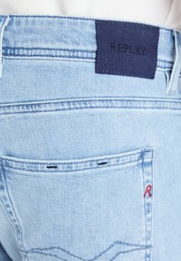 Světlé modré džíny z denimu s námořnicky modrým štítkem "REPLAY" na pásku, černým prošíváním, kapsou s červeným akcentem a měkkou texturou.