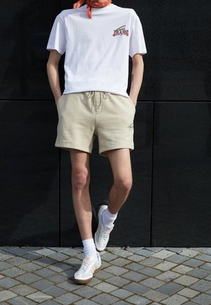 Biała koszulka graficzna z napisem "JEANS", beżowe spodenki z sznurkiem, oraz białe sneakersy z beżowymi akcentami. Model stoi na kamiennej nawierzchni.