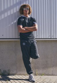 Sort atletisk skjorte med blå accenter, parret med sorte joggingbukser med logo. Båret med grå og hvide sneakers, indendørs i en minimalistisk urban indstilling.