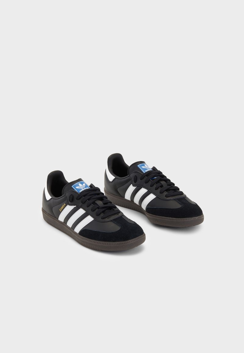 Een paar zwarte Adidas Samba sneakers met witte strepen, zwarte veters, gumzolen en een blauw Adidas-logo op de tong, tegen een witte achtergrond.