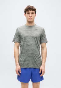 Γκρι αθλητική κοντομάνικη μπλούζα με ανάμεικτη υφή και μαύρο λογότυπο, σε συνδυασμό με μπλε σορτς, το μοντέλο στέκεται μπροστά σε απλό φόντο.