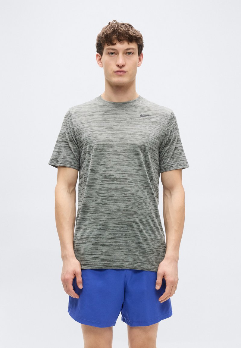 Γκρι αθλητική κοντομάνικη μπλούζα με ανάμεικτη υφή και μαύρο λογότυπο, σε συνδυασμό με μπλε σορτς, το μοντέλο στέκεται μπροστά σε απλό φόντο.