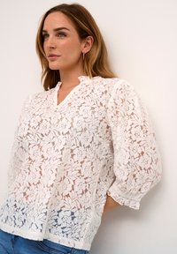 Blouse en dentelle blanche avec des motifs floraux, décolleté en V et manches bouffantes trois-quarts. Présente une coupe décontractée et un tissu texturé.