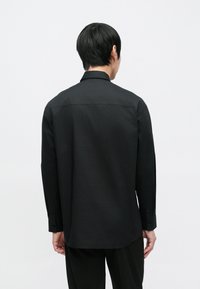 Crna košulja s dugim rukavima i ovratnikom na kopčanje, ravnog kroja, glatke teksture i prianjajućih manžeta, s nenametljivim dizajnom.