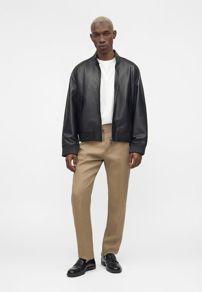 Άνδρας με κοντά ξανθά μαλλιά που φοράει μαύρο δερμάτινο μπουφάν, λευκό πουκάμισο, μπεζ παντελόνι και μαύρα παπούτσια, στέκεται μπροστά σε μονόχρωμο φόντο.