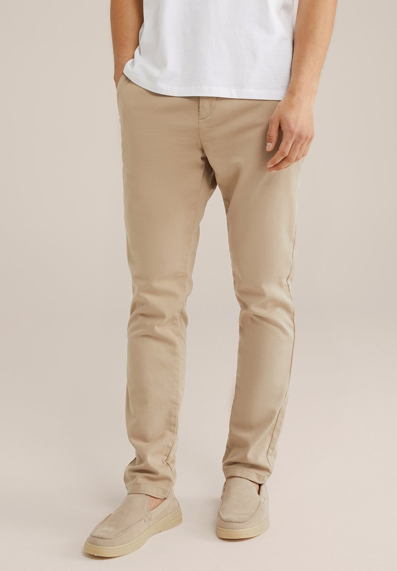 Pantalones de algodón beige con corte slim, frente plano, bolsillos laterales y dobladillos. Combinados con zapatos de slip-on beige claro.