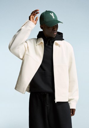 White textured jacket over a black hoodie, paired with a green cap featuring a white "Y." The model is raising a hand.