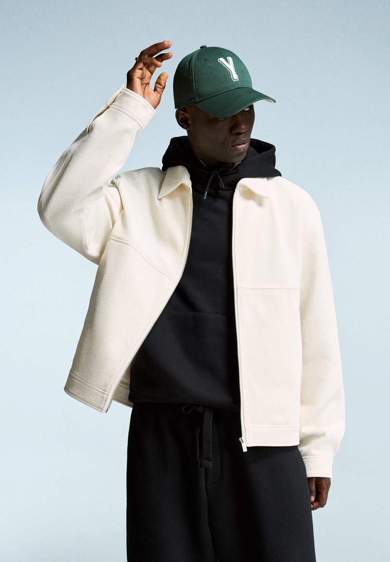White textured jacket over a black hoodie, paired with a green cap featuring a white "Y." The model is raising a hand.