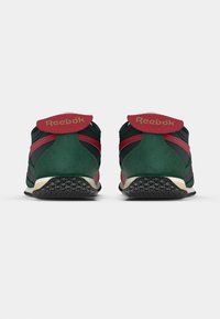 Um par de sapatilhas Reebok verdes e vermelhas visto por trás, com solas texturizadas pretas, em fundo branco.