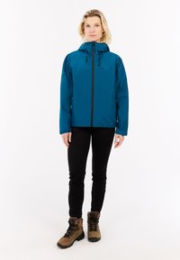 Tamsiai mėlyna nepralaidi vandeniui striukė su užtrauktuku ir reguliuojama kapu, derinama su juodomis kelnėmis ir rudos spalvos žygio batais, turinčiais raištelius ir tekstūruotus padus.