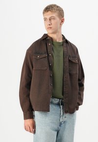 Young man wearing a dark brown button-up overshirt over a green ribbed shirt and light blue jeans, standing against a white background.