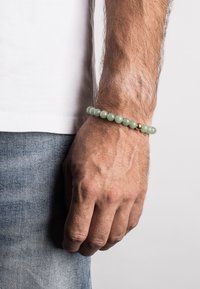 Bracciale di perline verdi con accenti dorati, indossato su un polso. Le perline sono lisce con una finitura lucida. Mano appoggiata su un tessuto in denim.
