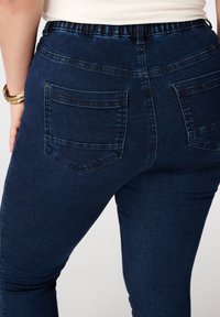 Donkerblauwe stretchdenim jeans met een hoge taille, elastische tailleband en twee achterzakken met horizontale stiksels.