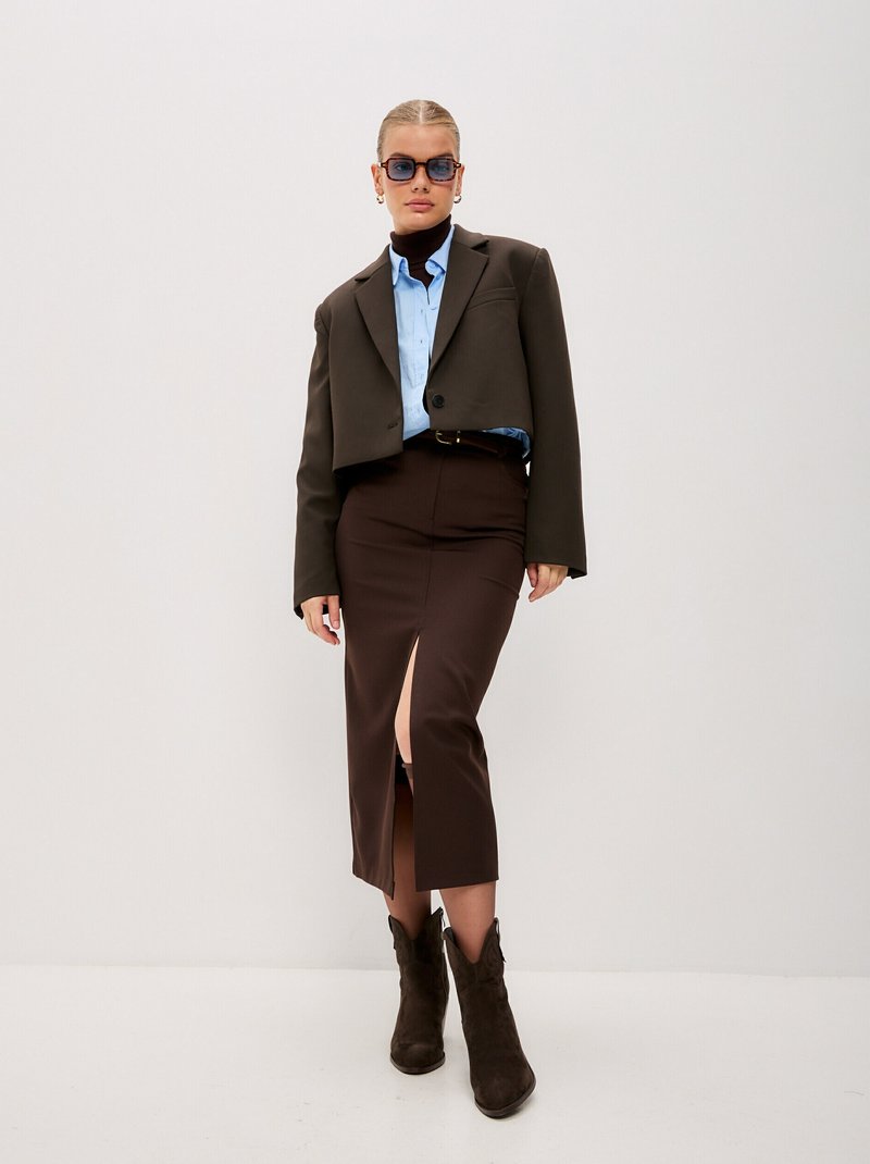 Kratší hnědý blazer, světlá modrá košile, hnědá sukně s bočním rozparkem a hnědé kovbojské boty. Čtvercové sluneční brýle celý outfit doplňují.