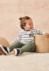 Kleinkind in einem weißen und marineblau gestreiften Langarmshirt mit Traktormotiv, grüner Hose und marineblauen Sneakers, das auf einer strukturierten Oberfläche sitzt.