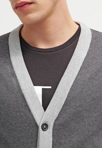 Graue Strickjacke mit hellgrauem, geripptem Kragen und einem einzelnen schwarzen Knopf, getragen über einem schwarzen Hemd mit weißem Logo.