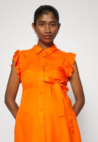 Vestido laranja com botões, com mangas em forma de rufos, detalhe de cintura com laço e textura suave. Apresenta fecho com botões e design de colarinho.