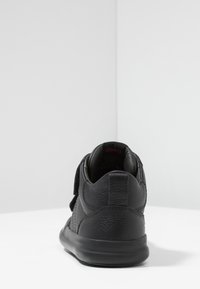 Sneaker in pelle nera con finitura testurizzata, dotato di collare imbottito, cinturino in Velcro e suola nera piatta. Vista posteriore su una superficie bianca.