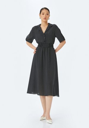 Schwarzes Kleid mit weißen Punkten, Kragen, kurzen Ärmeln und elastischem Bund. Luftiger, knielanger Rock aus leichtem Stoff.