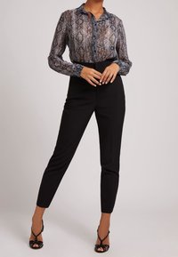 Blouse à imprimé serpent avec col, manches longues et matière transparente, associée à un pantalon noir taille haute et des talons noirs à lanières.