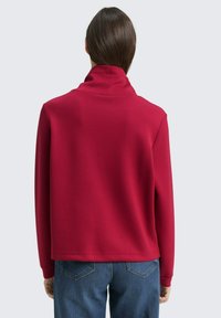Rotes Sweatshirt mit hohem Kragen, glatter Textur, langen Ärmeln und lockerem Schnitt, kombiniert mit blauen Jeans.