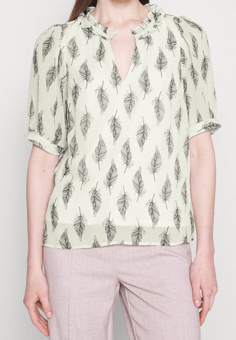 Blouse crème claire avec motif de plumes noires, col à volants, manches courtes et coupe décontractée, associée à un pantalon rose pâle.