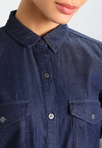 Camisa de denim oscuro con cuello puntiagudo, botones frontales y dos bolsillos en el pecho. La tela tiene una textura sutil y un color ligeramente deslavado.