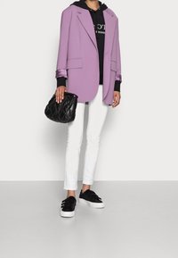 Blazer lilla, felpa nera con logo, jeans bianchi e borsa a tracolla nera trapuntata. Le scarpe slip-on nere con tre fasce completano il look.