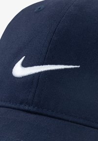Ljubičasta bejzbol kapa od pamuka, s bijelim vezenim Nike logotipom na prednjoj strani. Prikaz krupnog plana koji pokazuje teksturu i detalje šivanja.