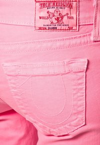 Jeans en denim rose avec une étiquette de marque, présentant un design de poche distinctif et des accents de couture, soulignant une texture lisse et une coupe ajustée.